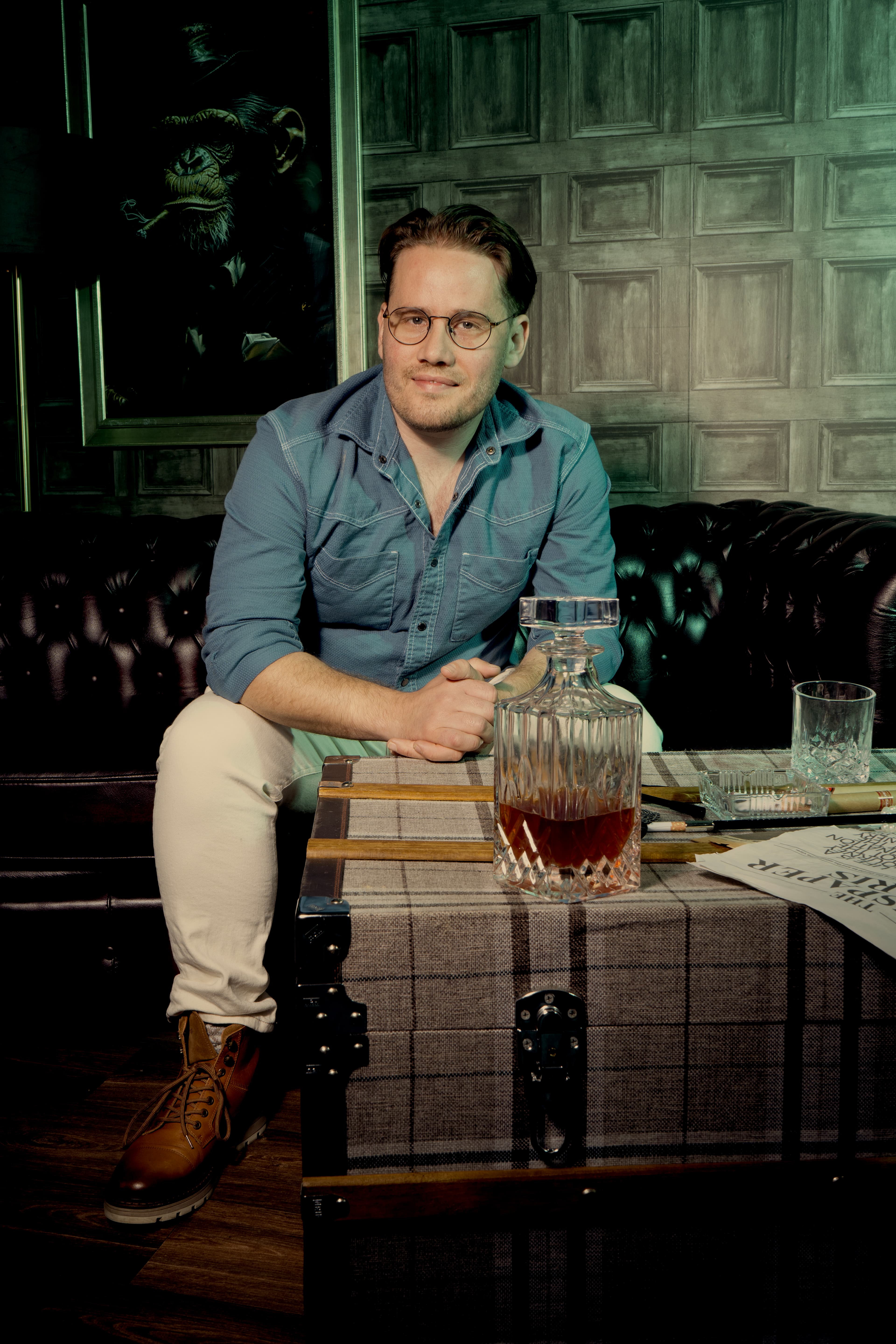 Man in glasses sits on a leather couch with a whiskey decanter on a trunk.